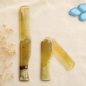 May include: Two folding combs made of translucent, mottled yellow and brown horn material. One comb is fully extended, while the other is partially folded, revealing a metal pivot pin. The combs feature fine teeth and a smooth, polished finish.