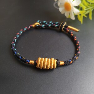 May include: A braided bracelet featuring a black cord interwoven with thin strands of blue, orange, and purple thread. The bracelet includes a central cylindrical gold-colored metallic bead with a ribbed texture and a sliding gold-colored knot closure.