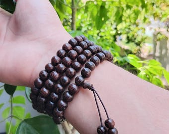 Op de afbeelding: Een donkerbruine houten kralenarmband is om een pols gewikkeld. De armband is gemaakt van meerdere strengen ronde kralen. De kralen hebben een rijke, donkerbruine kleur. De armband is vastgebonden met een donkerbruin touwtje.