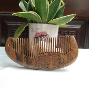 May include: A wooden comb with a curved handle and fine teeth. The comb is a dark brown colour with a decorative carved design on the handle. A small hole is drilled near the top of the handle. The comb is on a white surface.