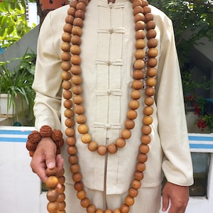 May include: A long necklace made of smooth, round, light brown wooden beads draped around the neck, paired with a matching wooden bead bracelet and a separate strand of beads held in the hand, worn over a beige long-sleeved shirt with toggle buttons.