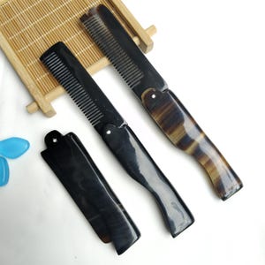 Puede incluir: Se muestran tres peines plegables con mangos y dientes de color marrón oscuro y negro. Un peine está abierto, revelando los dientes, mientras que los otros están cerrados. Los mangos tienen un aspecto veteado. Los peines están sobre una superficie blanca.