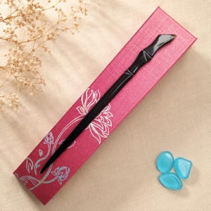 May include: A long, slender, black hair stick with a smooth, polished finish and a slightly curved, tapered end. It is positioned diagonally across a rectangular, magenta-colored gift box decorated with a white, linear floral pattern.