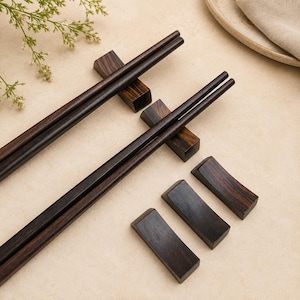 Puede incluir: Dos pares de palillos de madera de color marrón oscuro sobre soportes rectangulares de madera a juego, acompañados de tres soportes individuales adicionales de madera marrón oscuro, todos con un patrón de veta de madera natural visible.