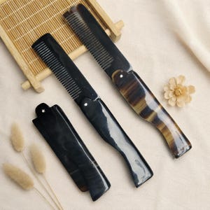 May include: Three folding combs made of polished dark horn with natural brown and black marbled patterns. Two combs are fully extended, showing fine teeth and a curved handle, while one comb is folded shut into a compact, smooth rectangular shape.