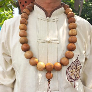 May include: A long necklace featuring large, round, light brown wooden beads with visible grain patterns. The necklace includes a central bead with a smaller dangling pendant consisting of two smaller beads on a thin brown cord, worn over a beige shirt.