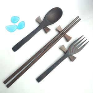 Puede incluir: Un juego de cubiertos de madera marrón oscuro que incluye una cuchara, palillos y un tenedor, cada uno apoyado en un pequeño soporte de madera. Los utensilios se muestran sobre un fondo blanco, con tres guijarros de vidrio azul. La cuchara, los palillos y el tenedor son de madera oscura.