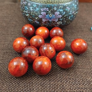 May include: A selection of reddish-brown wooden beads with a polished surface. Each bead is spherical and has a small hole. A decorative ceramic bowl with floral patterns is in the background.
