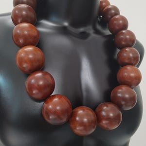 May include: A chunky statement necklace composed of large, spherical, polished dark brown wooden beads. The beads are uniform in shape and size, strung together to form a continuous circular collar that rests around the neck area.