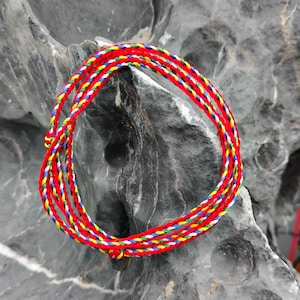 Può includere: Un braccialetto a corda rosso arrotolato con fili gialli, verdi, blu e bianchi. Il braccialetto è realizzato con fili fittamente intrecciati ed è esposto su una superficie di pietra grigia strutturata. Un accessorio vibrante.