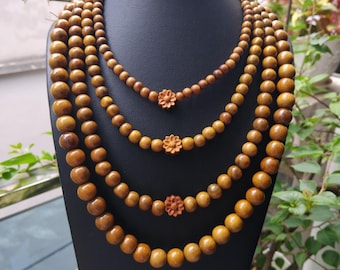 Puede incluir: Un collar de múltiples hilos con cuatro capas graduadas de cuentas de madera redondas, lisas y de color marrón claro. Cada hilo está centrado con una pequeña cuenta de madera tallada en forma de flor. El collar se exhibe en un busto negro.