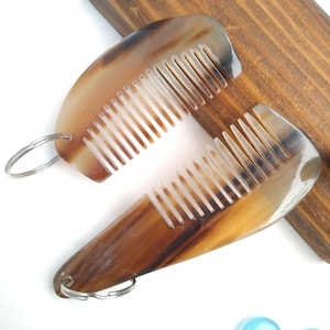 Puede incluir: Dos peines de cuerno con un patrón marrón y beige natural. Cada peine tiene una fila de dientes y un anillo de metal plateado. Los peines se muestran sobre una superficie de madera.