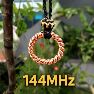 May include: A copper-colored metal ring with a twisted texture, suspended by a black braided cord featuring a decorative black and gold knot accent. The text 144MHz is displayed in yellow at the bottom of the image, indicating a specific frequency.