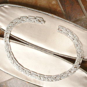 May include: A silver-toned metal necklace featuring a double-row chain design composed of interconnected geometric links. Each link is encrusted with small, clear, sparkling rhinestones that catch the light, creating a continuous, shimmering effect.