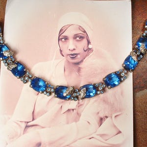 Pode incluir: Pulseira vintage com grandes strass azuis ovais e redondos. A pulseira tem uma moldura de metal prateado e detalhes de folhas. Apresentada numa fotografia vintage de uma mulher.