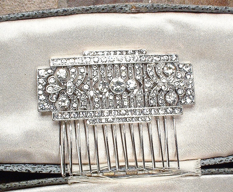 May include: A silver hair comb with a decorative design of clear rhinestones. The comb has a geometric pattern with a central floral motif.