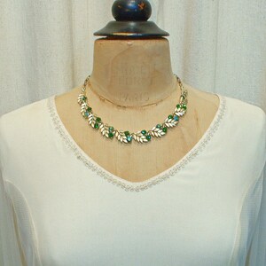 May include: A gold-toned necklace with a leaf and gemstone design. The necklace features white leaves with green and blue gemstones. The necklace is displayed on a vintage mannequin with the text "MODELE ET FORET PARIS".