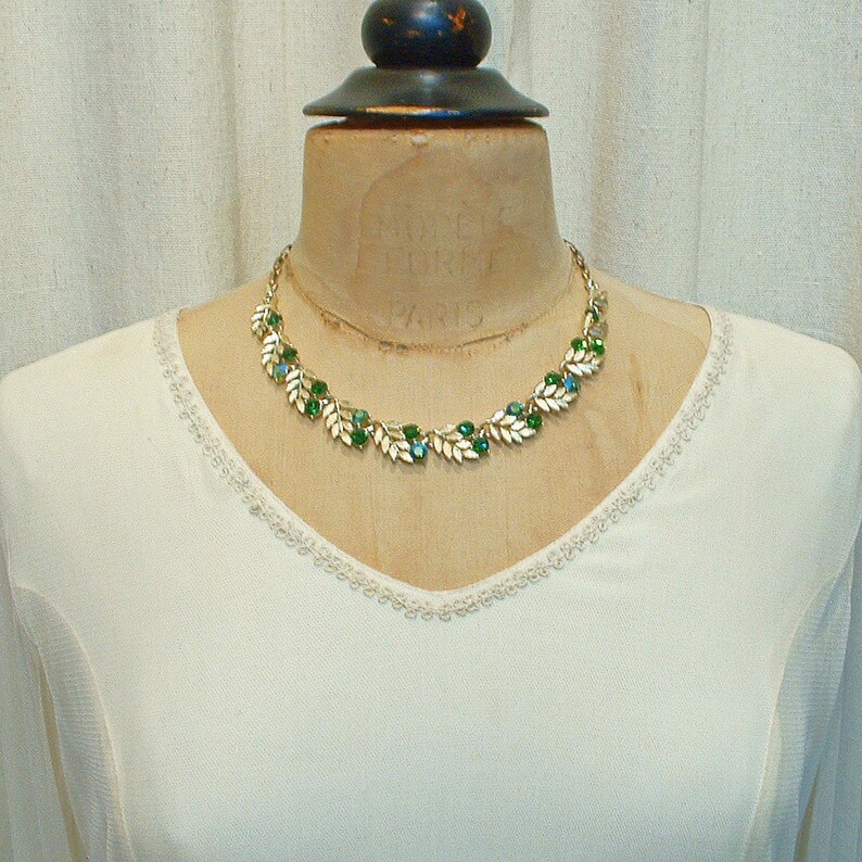 May include: A gold-toned necklace with a leaf and gemstone design. The necklace features white leaves with green and blue gemstones. The necklace is displayed on a vintage mannequin with the text "MODELE ET FORET PARIS".