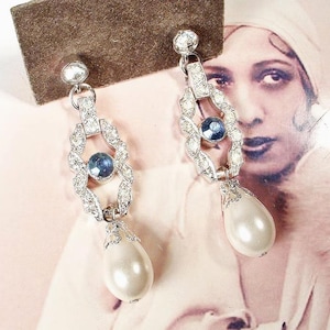 Peut inclure: Boucles d'oreilles Art Déco argentées avec des pierres précieuses claires et bleues et une goutte de perle en forme de poire. Les boucles d'oreilles sont présentées sur une surface marron.