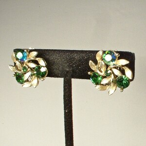 May include: A pair of vintage clip-on earrings. Each earring features a cluster of green and iridescent blue-green gemstones, surrounded by gold-toned leaf accents. The earrings are displayed on a black stand.