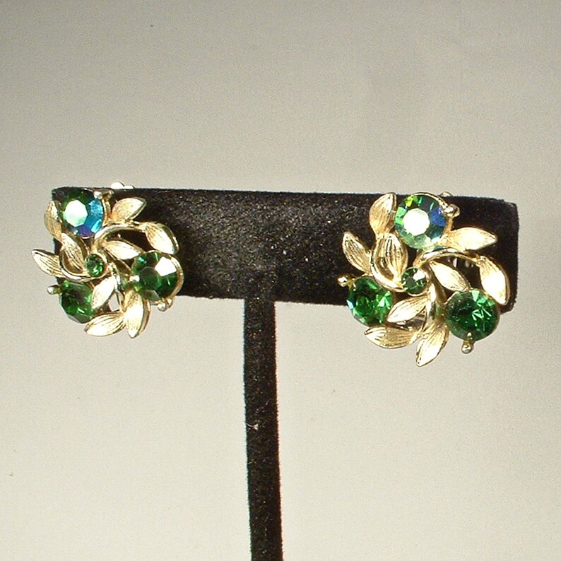 May include: A pair of vintage clip-on earrings. Each earring features a cluster of green and iridescent blue-green gemstones, surrounded by gold-toned leaf accents. The earrings are displayed on a black stand.