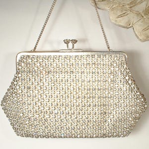 May include: A silver-toned clutch handbag covered in clear, faceted rhinestones arranged in a grid pattern. The bag features a metallic frame, a kiss-lock closure with two circular knobs, and a thin, detachable silver-toned chain shoulder strap.