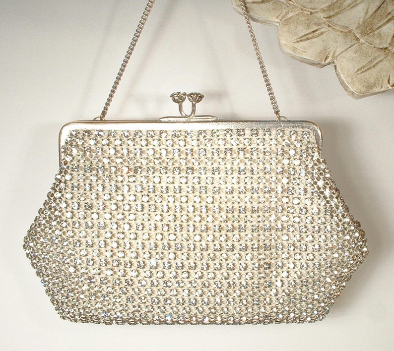 May include: A silver-toned clutch handbag covered in clear, faceted rhinestones arranged in a grid pattern. The bag features a metallic frame, a kiss-lock closure with two circular knobs, and a thin, detachable silver-toned chain shoulder strap.