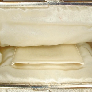 May include: An open evening bag with a silver-toned metal frame and a smooth, cream-colored satin interior lining. The interior features a single slip pocket centered on the back wall. The exterior edges are adorned with clear, sparkling crystals.