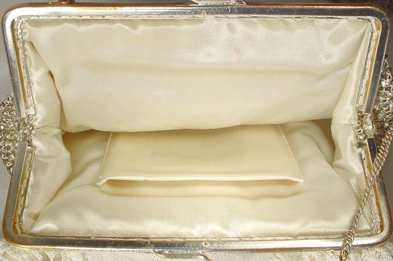 May include: An open evening bag with a silver-toned metal frame and a smooth, cream-colored satin interior lining. The interior features a single slip pocket centered on the back wall. The exterior edges are adorned with clear, sparkling crystals.