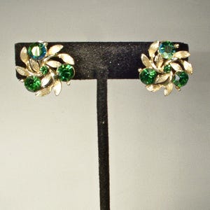 May include: A pair of vintage clip-on earrings. Each earring features a cluster of green and teal-colored rhinestones surrounded by gold-toned leaves. The earrings are displayed on a black stand against a neutral background.
