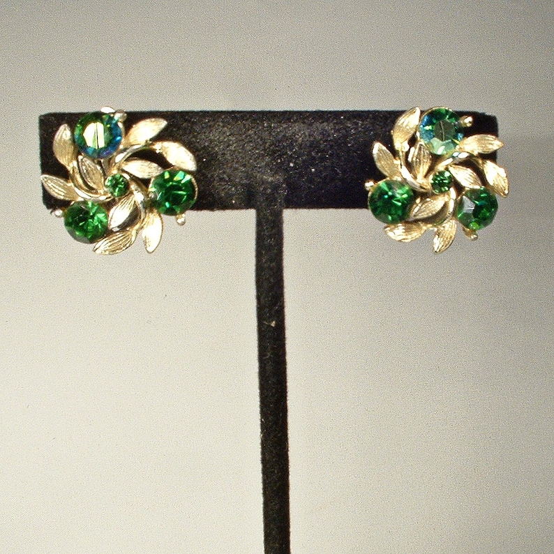 May include: A pair of vintage clip-on earrings. Each earring features a cluster of green and teal-colored rhinestones surrounded by gold-toned leaves. The earrings are displayed on a black stand against a neutral background.