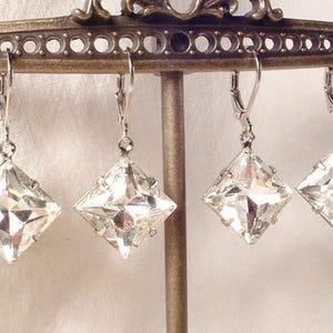 Puede incluir: Cuatro pendientes de cristal transparente, tallados en forma cuadrada, con cierres de palanca plateados, cuelgan de una exhibici&oacute;n met&aacute;lica decorativa. Los pendientes est&aacute;n facetados, reflejando la luz. El soporte de exhibici&oacute;n es de color bronce.
