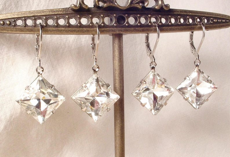 Puede incluir: Cuatro pendientes de cristal transparente, tallados en forma cuadrada, con cierres de palanca plateados, cuelgan de una exhibici&oacute;n met&aacute;lica decorativa. Los pendientes est&aacute;n facetados, reflejando la luz. El soporte de exhibici&oacute;n es de color bronce.