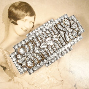 May include: A silver-toned, geometric brooch with a floral design, set with clear rhinestones.