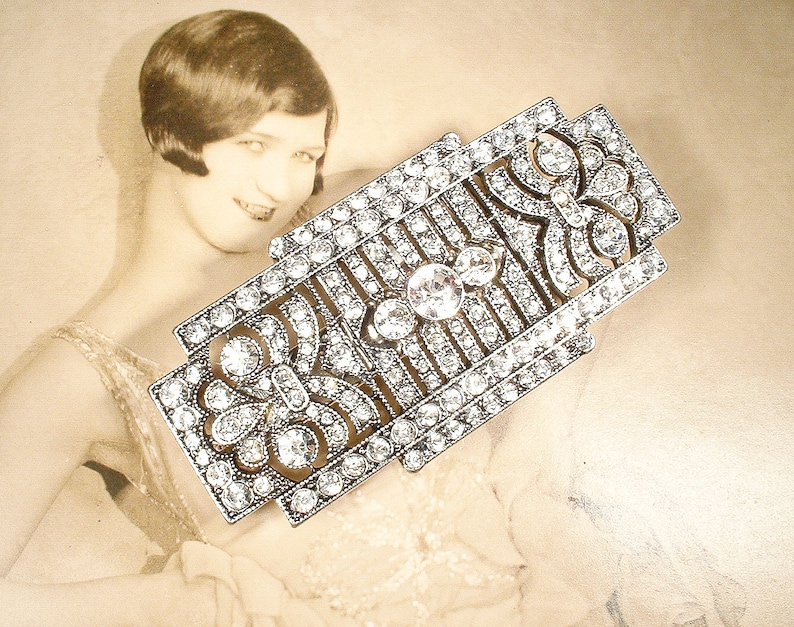 May include: A silver-toned, geometric brooch with a floral design, set with clear rhinestones.