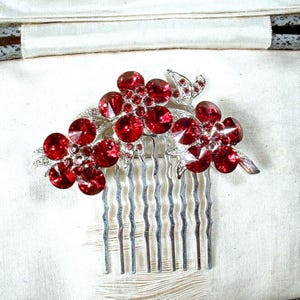 Könnte beinhalten: Ein silberfarbener Haarkamm mit einem dekorativen Blumenarrangement, das mit zahlreichen runden, facettierten roten Kristallen besetzt ist. Der Metallkamm hat sieben gewellte Zinken und ist mit metallischen Blättern zwischen den roten Blüten verziert.