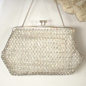 May include: A silver-toned evening handbag completely covered in clear, sparkling rhinestones. The bag features a structured frame, a kiss-lock closure with two small decorative knobs, and a thin, delicate silver-toned metal chain shoulder strap.
