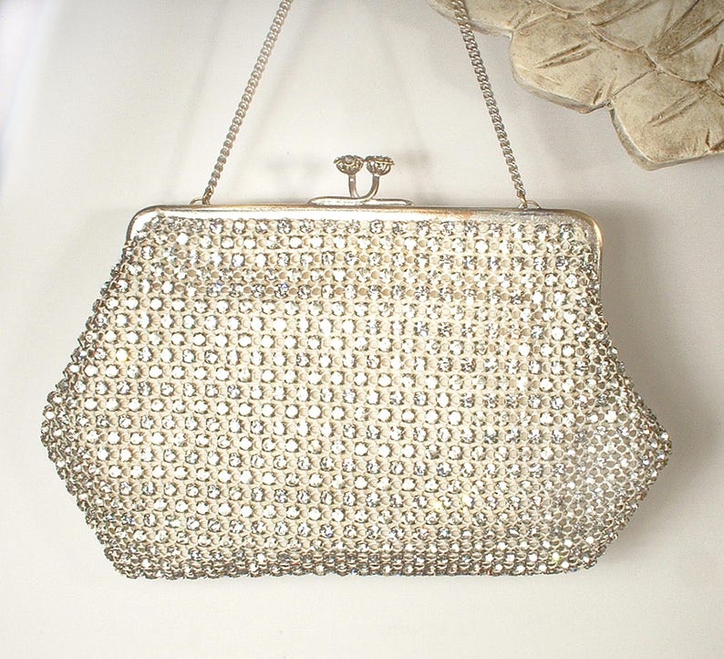 May include: A silver-toned evening handbag completely covered in clear, sparkling rhinestones. The bag features a structured frame, a kiss-lock closure with two small decorative knobs, and a thin, delicate silver-toned metal chain shoulder strap.