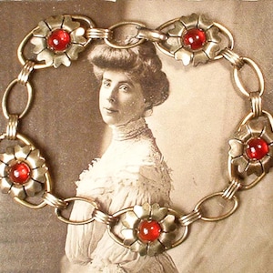 Puede incluir: Un brazalete de cadena dorada con cinco acentos de flores de vidrio rojo. El brazalete está hecho de cadena de eslabones con un diseño de flores decorativo.