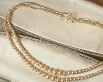 May include: A vintage pearl necklace with two strands of varying pearl sizes. The necklace is fastened with a decorative clasp featuring small pearls and clear crystals. The necklace rests on a cream-colored satin surface inside a vintage jewelry box.