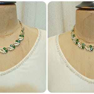 May include: A gold-toned necklace with green and clear gemstones and leaf-shaped accents. The necklace is displayed on a mannequin, set against a white top with a decorative neckline. The jewelry is a vintage style.