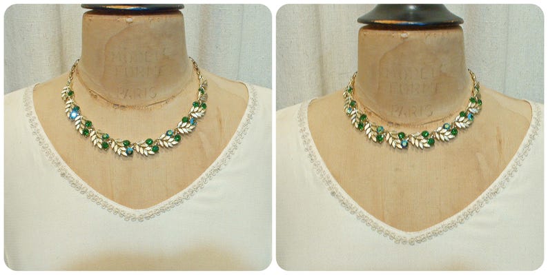 May include: A gold-toned necklace with green and clear gemstones and leaf-shaped accents. The necklace is displayed on a mannequin, set against a white top with a decorative neckline. The jewelry is a vintage style.