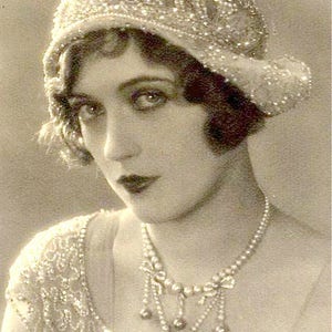Puede incluir: Retrato en sepia de una mujer con un sombrero cloche con cuentas y un collar de perlas. El sombrero y el vestido est&aacute;n adornados con intrincados abalorios. El texto "Amore Treasure" est&aacute; escrito en cursiva en la parte inferior de la imagen.