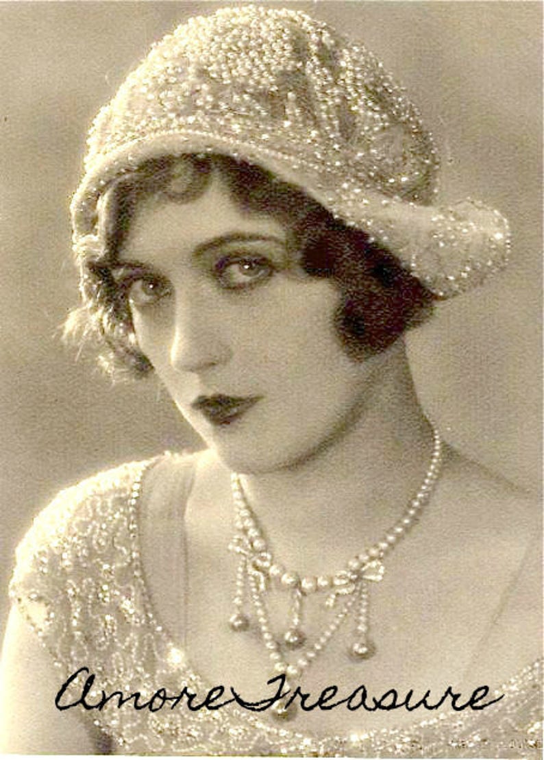 Puede incluir: Retrato en sepia de una mujer con un sombrero cloche con cuentas y un collar de perlas. El sombrero y el vestido est&aacute;n adornados con intrincados abalorios. El texto "Amore Treasure" est&aacute; escrito en cursiva en la parte inferior de la imagen.