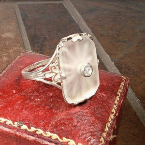 Op de afbeelding: Een zilveren ring met een heldere, rechthoekige steen gezet in een filigraan ontwerp. De steen heeft een kleinere, heldere, diamantvormige steen in het midden.