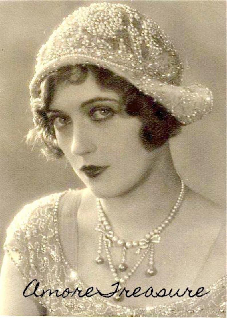 Pu&ograve; includere: Un ritratto in bianco e nero di una donna che indossa un cappello e una collana di perle. La donna guarda la fotocamera e ha un'espressione seria sul viso. Il testo "Amore Treasure" &egrave; scritto in corsivo nella parte inferiore dell'immagine.