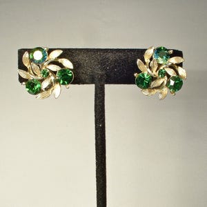 May include: A pair of gold-toned clip-on earrings. Each earring features a cluster of green and teal-colored gemstones surrounded by gold-colored leaf accents. The earrings are displayed on a black stand.