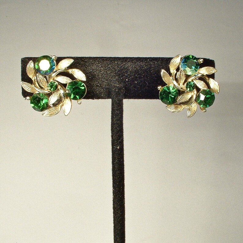 May include: A pair of gold-toned clip-on earrings. Each earring features a cluster of green and teal-colored gemstones surrounded by gold-colored leaf accents. The earrings are displayed on a black stand.
