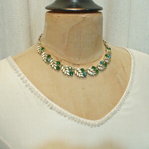 May include: A gold-toned necklace with leaf-shaped accents and green and turquoise gemstones. The necklace is displayed on a mannequin wearing a cream-colored top.