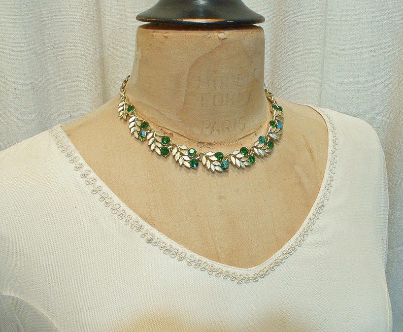 May include: A gold-toned necklace with leaf-shaped accents and green and turquoise gemstones. The necklace is displayed on a mannequin wearing a cream-colored top.